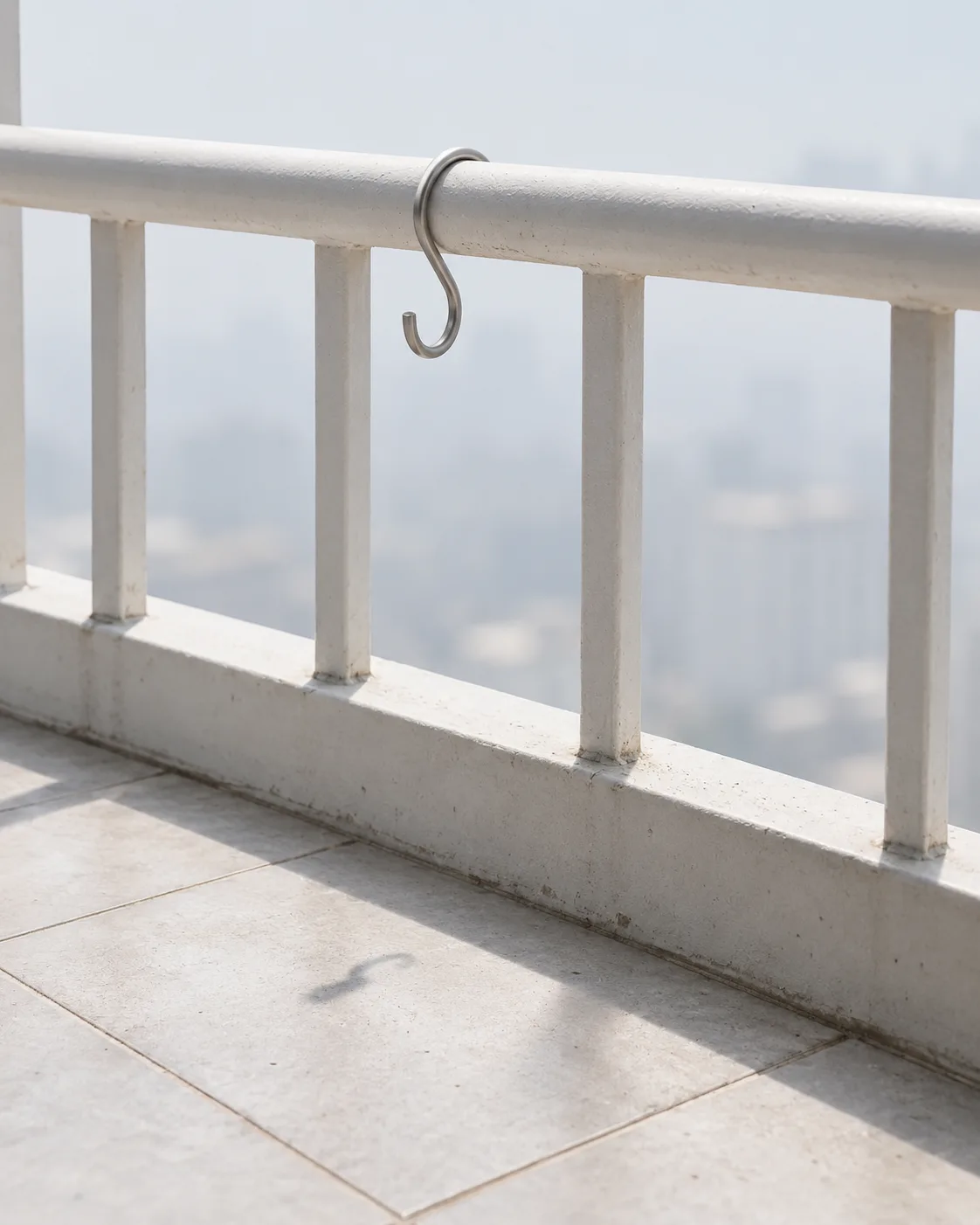 An empty steel S-hook hangs from a white balcony railing at midday in summer, its soft shadow on pale grey tiles, a hazy city beyond.